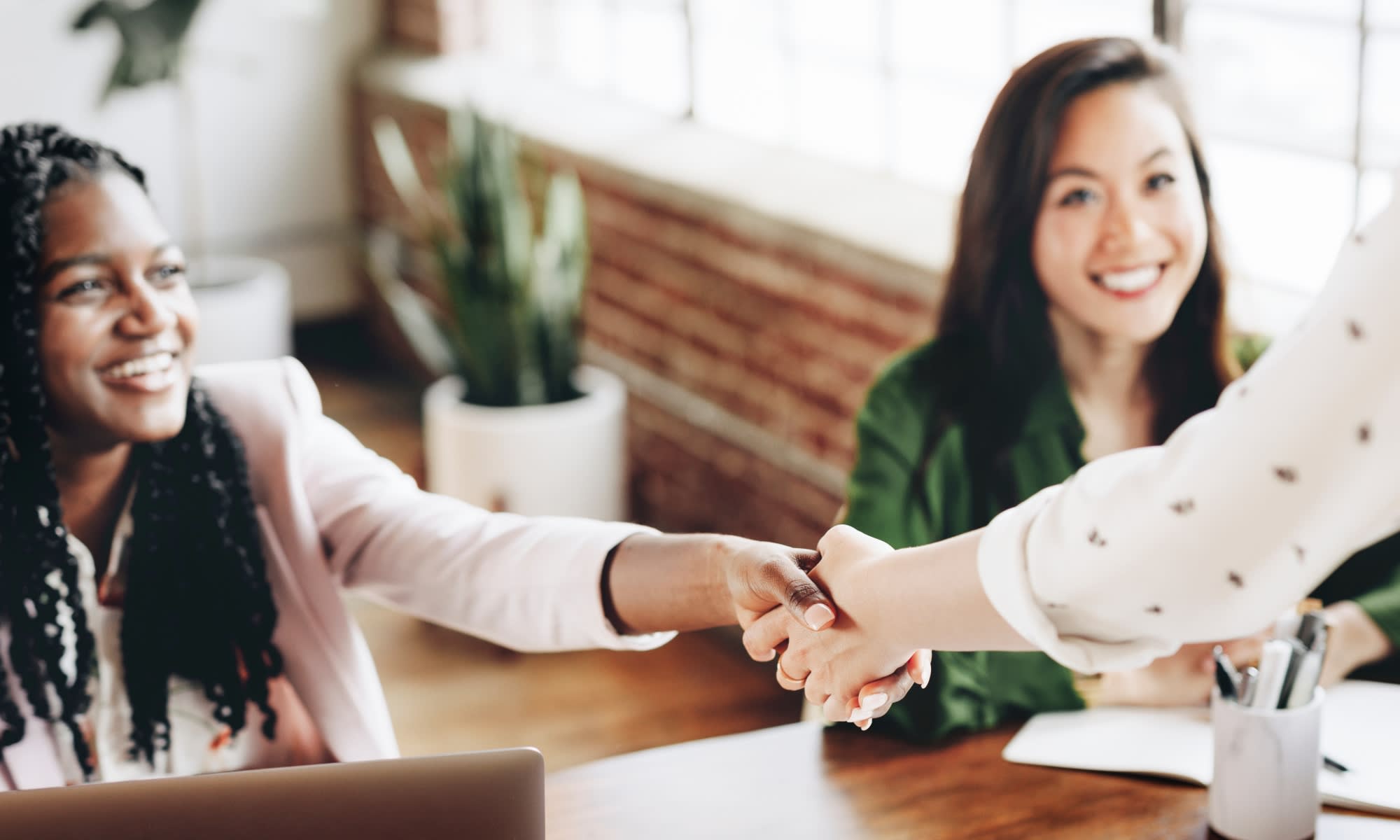 Close up of smiling professional woman in an office shaking hands with another person out of the picture with another woman looking on