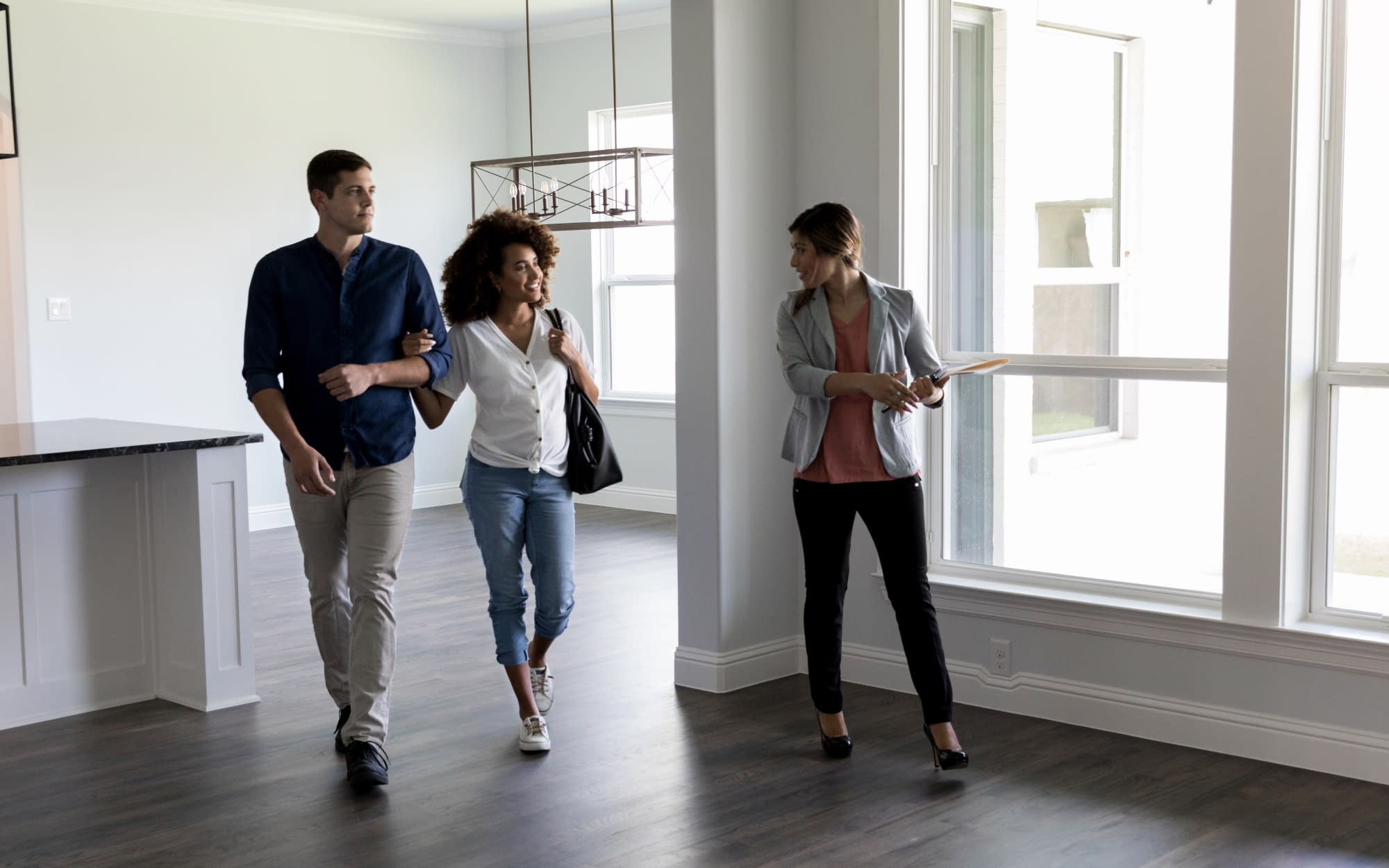 Young real estate agent looking at young couple as they walk through empty home for sale with hardwood floors and light coming through large windows